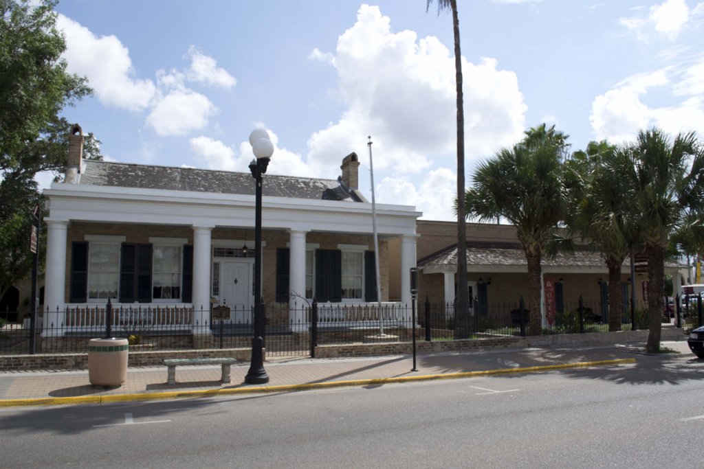 Historic white colonial-style building with columns and black shutters alongside a tan brick building, palm trees, and wrought iron fencing on a sunny Texas street